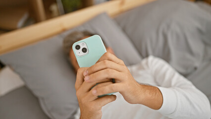Young, handsome hispanic man with grey hair, comfortably chilling in bed, engrossed in texting on...