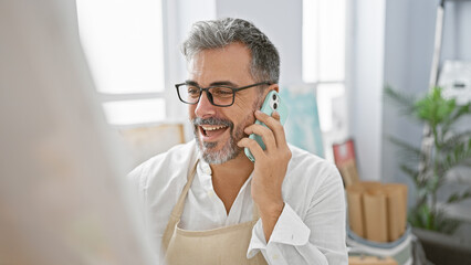 Handsome young hispanic artist with grey hair, engrossed in a vibrant phone conversation while confidently drawing at his bustling art studio.