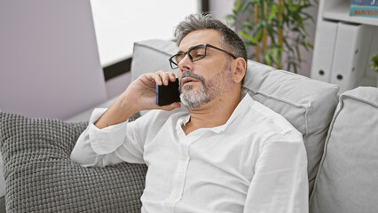 Exhausted young hispanic man with grey hair, engrossed in a serious phone conversation, slumped on a living room sofa at home