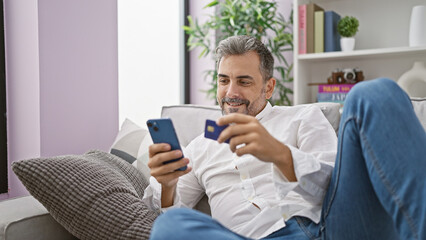 Smiling young grey-haired hispanic man casually shopping online, safely paying with credit card on sofa at home, enjoying technology