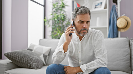 Fototapeta premium Young hispanic grey-haired man speaking on the phone sitting on sofa at home