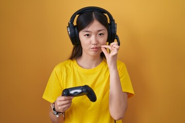 Chinese young woman playing video game holding controller mouth and lips shut as zip with fingers....