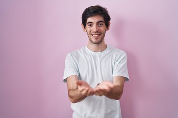 Young hispanic man standing over pink background smiling with hands palms together receiving or giving gesture. hold and protection