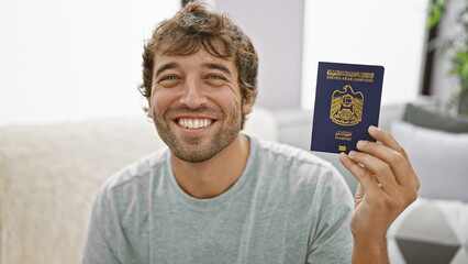 Chilled-out young man, smiling happily with confidence, casually sitting on sofa at home, holding his travel passport of united arab emirates, ready to enjoy his fun vacation trip - obrazy, fototapety, plakaty