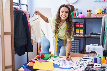 Fototapeta premium Young beautiful hispanic woman tailor smiling confident holding t shirt writing on notebook at clothing factory
