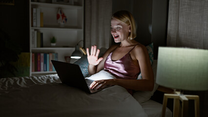 Young blonde woman doing video call sitting on the bed at bedroom