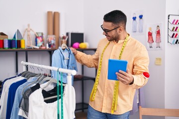 Young hispanic man tailor holding clothes on rack using touchpad at tailor shop