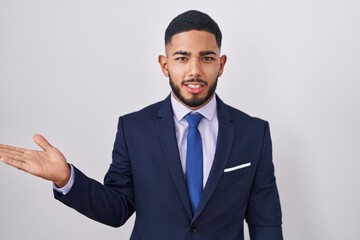 Young hispanic man wearing business suit and tie smiling cheerful presenting and pointing with palm of hand looking at the camera.