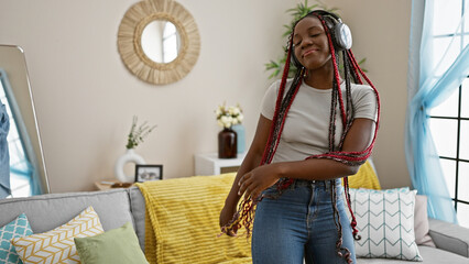 Joyous african american woman rocking braids, listening to music, dancing with confidence in the living room, smiling as she enjoys her relaxing lifestyle at home