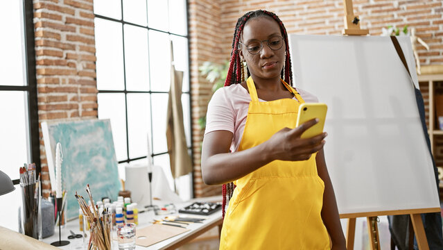 Beautiful african-american woman artist paints her canvas masterpiece, standing in apron with her smartphone at a learning center art studio