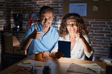 Middle age hispanic couple using touchpad sitting on the table at night doing happy thumbs up gesture with hand. approving expression looking at the camera showing success.