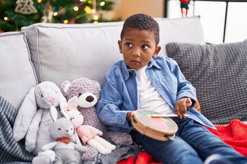 African american boy playing tambourine sitting on sofa by christmas tree at home - obrazy, fototapety, plakaty