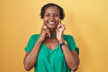 African woman with dreadlocks standing over yellow background smiling with open mouth, fingers...