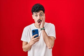 Hispanic man using smartphone over red background afraid and shocked, surprise and amazed expression with hands on face