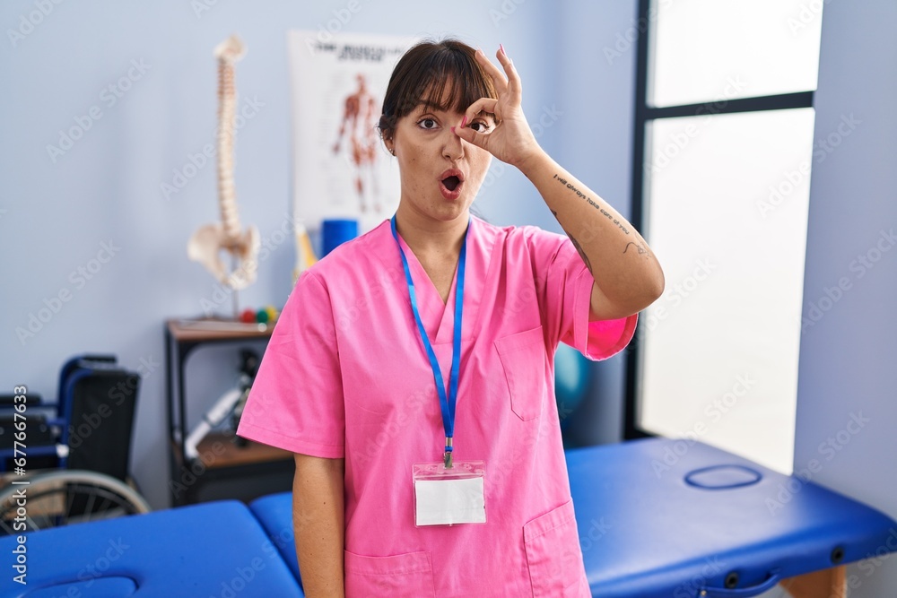 Canvas Prints Young brunette woman working at rehabilitation clinic doing ok gesture shocked with surprised face, eye looking through fingers. unbelieving expression.