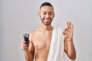 Young hispanic man holding electric razor doing ok sign with fingers, smiling friendly gesturing excellent symbol - obrazy, fototapety, plakaty