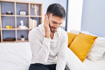 Young hispanic man suffering for ear pain sitting on bed at bedroom