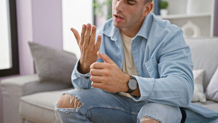 Unhappy, young hispanic man reluctantly sitting in his cozy living room, achingly nursing finger...