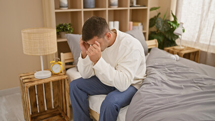 Fototapeta premium Exhausted young hispanic man stressed and worried, sitting on bed in his bedroom, struggling with problems and anxiety