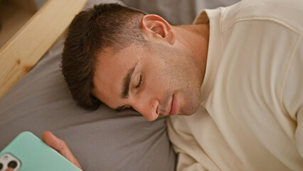 Exhausted young hispanic man, holding his phone, deeply asleep, resting comfortably in a cozy bed...