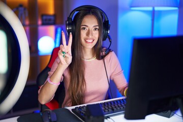 Young hispanic woman playing video games showing and pointing up with fingers number two while smiling confident and happy. - obrazy, fototapety, plakaty