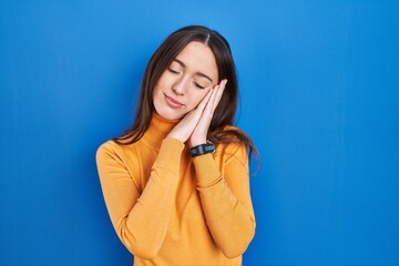 Young brunette woman standing over blue background sleeping tired dreaming and posing with hands together while smiling with closed eyes.