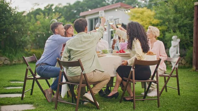 Group of several multiethnic young men and women are sitting at table outdoors at home backyard, eating, communicating and clinking champagne glasses. Celebration, friendship and holidays concept.
