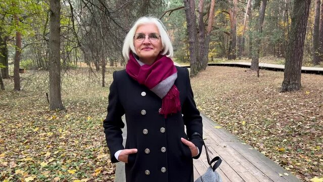 Portrait of attractive elderly 60 year old woman with gray hair and eyeglasses in nature forest or park looks at camera and smiles. Pretty Senior female model in warm clothes outdoors.