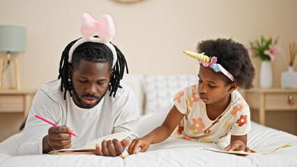 African american father and daughter wearing funny diadem drawing on notebook lying on bed at...
