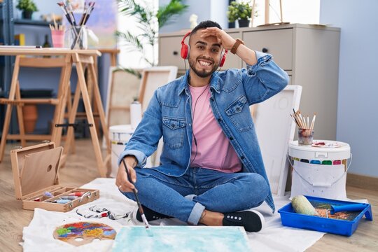 Young hispanic man painter sitting on the floor at art studio stressed and frustrated with hand on head, surprised and angry face