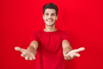 Young hispanic man standing over red background smiling cheerful offering hands giving assistance...