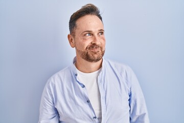 Middle age caucasian man standing over blue background smiling looking to the side and staring away thinking.