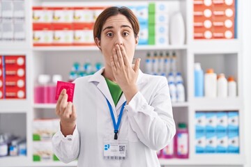 Brunette woman working at pharmacy drugstore holding condom covering mouth with hand, shocked and...