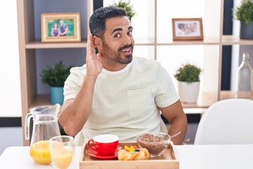 Fototapeta na wymiar Hispanic man with beard eating breakfast smiling with hand over ear listening and hearing to rumor or gossip. deafness concept.