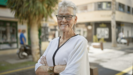 Confident grey-haired senior woman enjoying street life! smiling, arms crossed, she exudes casual confidence amid urban sunshine. looking cheery, she's a beacon of mature happiness out there.