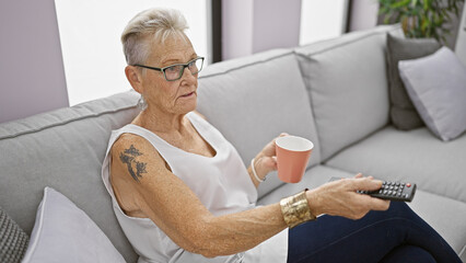 Serene grey-haired senior woman relaxing on a comfy sofa, deeply engrossed in a morning movie,...