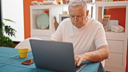 Middle age grey-haired man using laptop with relaxed expression at dinning room