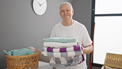 Middle age grey-haired man smiling confident holding folded towels at laundry room