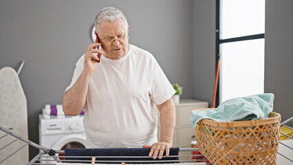 Middle age grey-haired man talking on smartphone hanging clothes on clothesline at laundry room