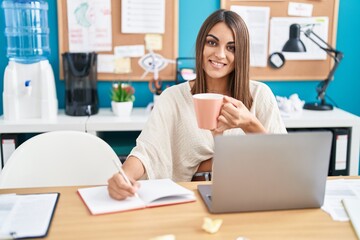Young beautiful hispanic woman business worker writing on notebook drinking coffee at office