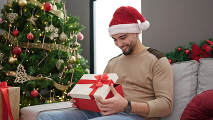 Young arab man unpacking gift sitting on sofa by christmas tree at home
