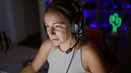 Attractive young blonde woman streamer, locked in her gaming room, focused and serious, using...