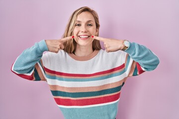 Young blonde woman standing over pink background smiling cheerful showing and pointing with fingers teeth and mouth. dental health concept.