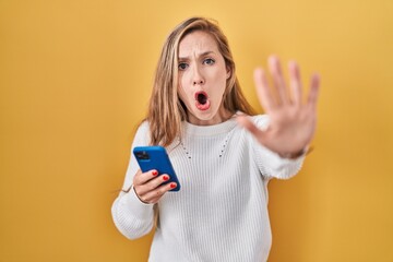 Young blonde woman using smartphone typing message doing stop gesture with hands palms, angry and...