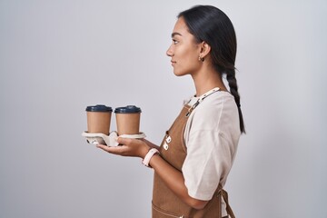 Young hispanic woman wearing professional waitress apron holding coffee looking to side, relax...