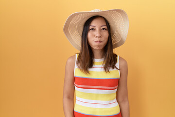 Middle age chinese woman wearing summer hat over yellow background puffing cheeks with funny face. mouth inflated with air, crazy expression. - obrazy, fototapety, plakaty