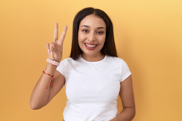 Young arab woman wearing casual white t shirt over yellow background showing and pointing up with fingers number two while smiling confident and happy.