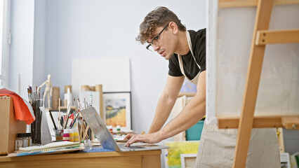 Attractive young hispanic man, a serious artist, standing by the easel in a well-lit art studio, passionately drawing on his laptop, immersed in the online creative sphere