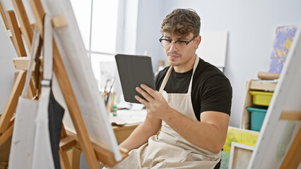 Attractive young hispanic man artist enthralled in drawing lesson using touchpad at art studio, seated indoors with brush in hand, canvas at easel & wearing glasses