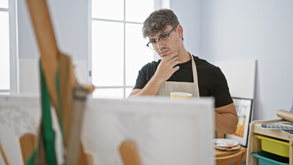 Determined young hispanic man in apron, an artist deep in thought, drinking coffee while looking at his drawing in art studio